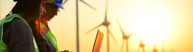 2 people looking at a laptop outside in front of wind turbines